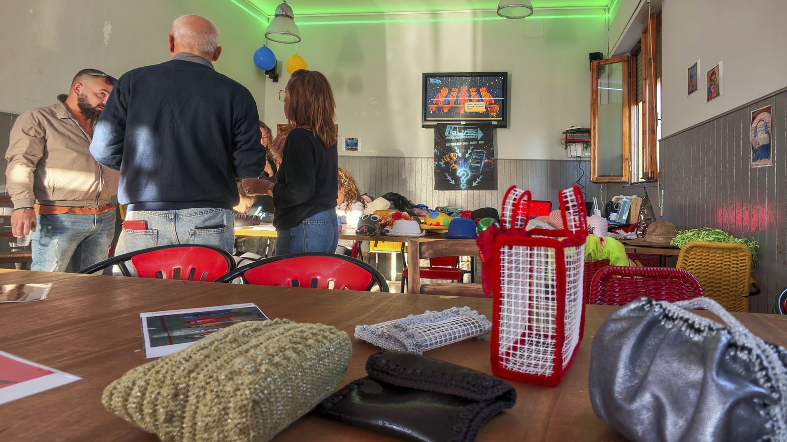 Persone che chiacchierano in sala al Circolo ARCI Gori durante l’Open Day di No Limits APS, con borse all’uncinetto di riciclo sul tavolo in primo piano